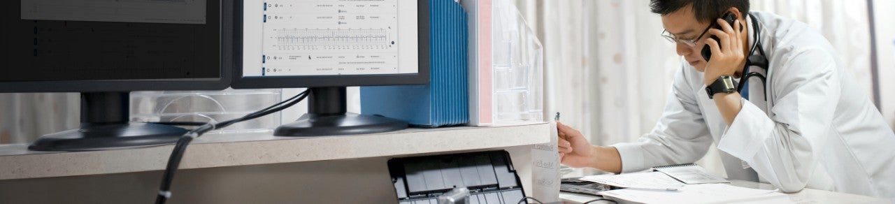 Healthcare provider on phone looking at patient files, as computer monitor displays cardiac data in foreground.