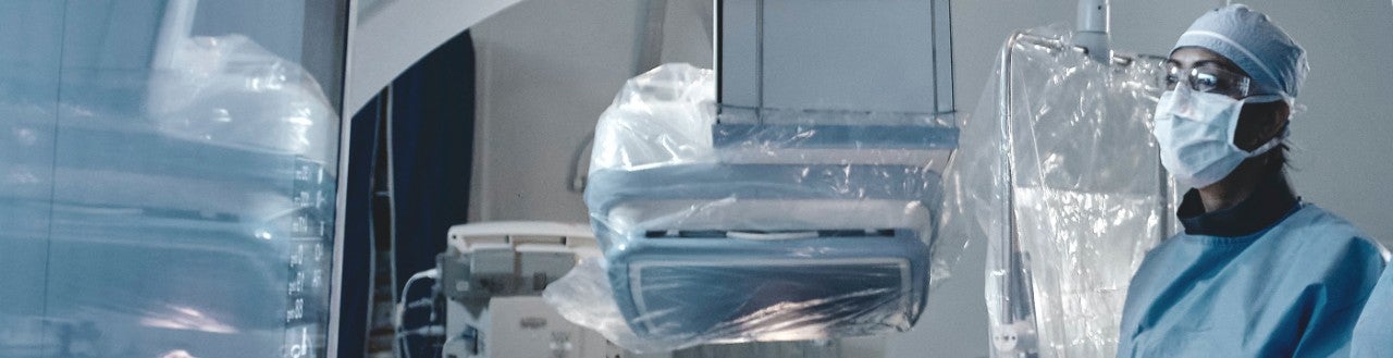 Doctor dressed in scrubs, mask and safety glasses, looks across surgical suite.