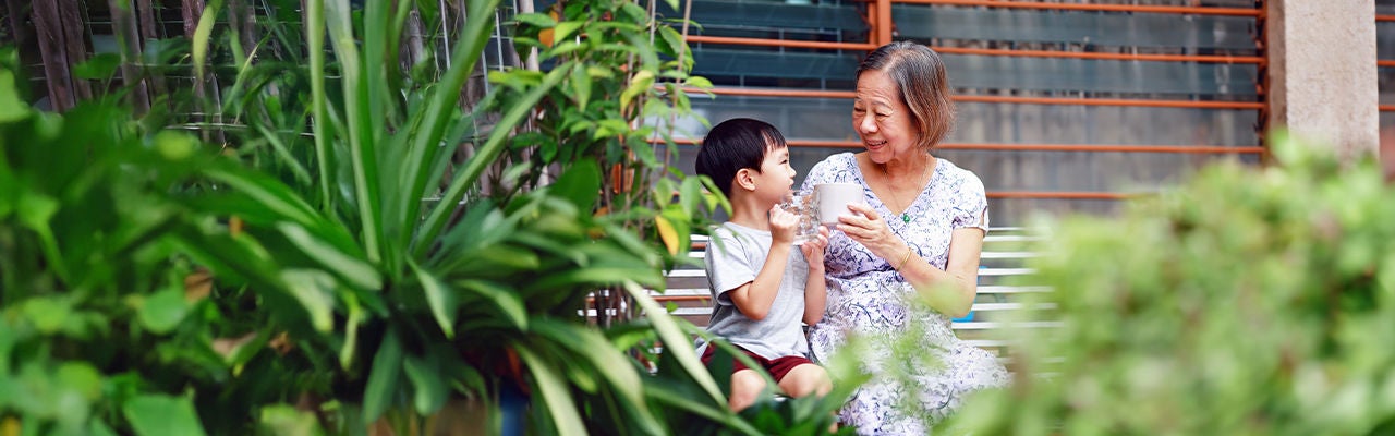 An older woman and a young child sit together on a bench in a lush garden setting.