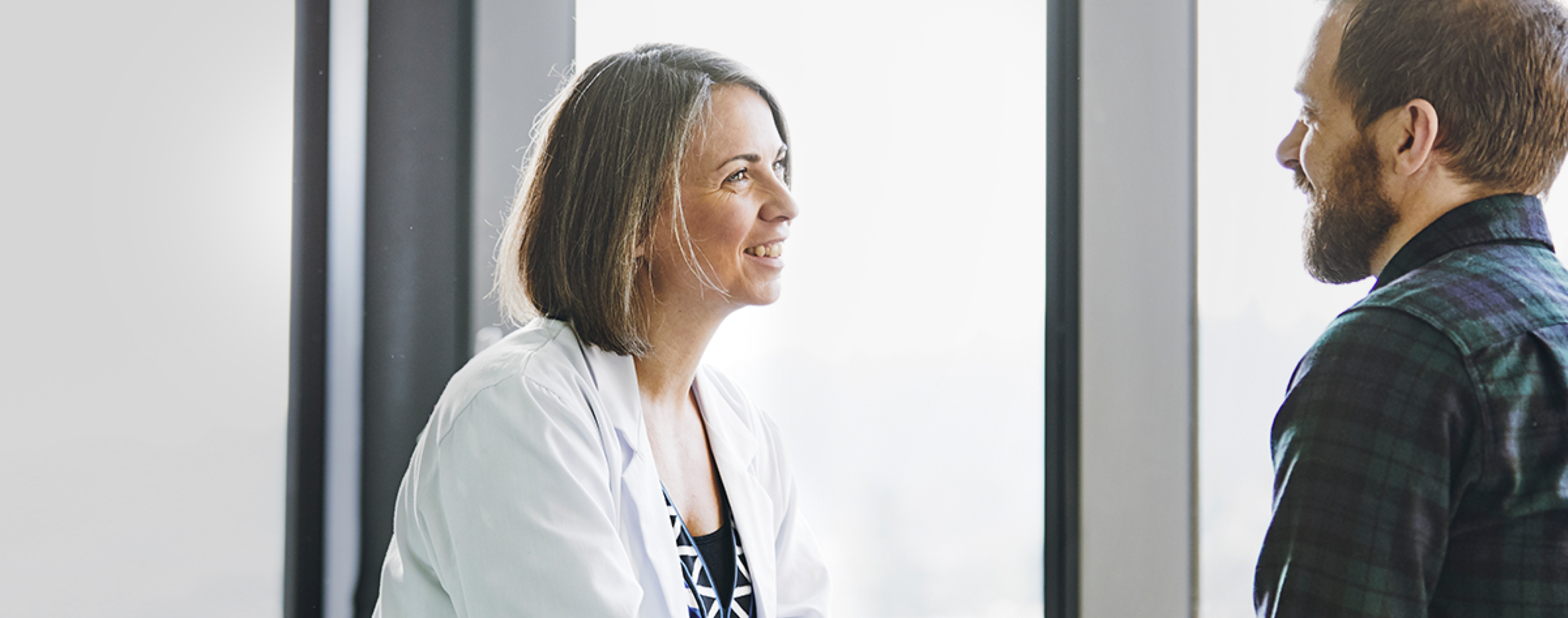 Smiling physician in white coat looking at smiling patient in plaid shirt.