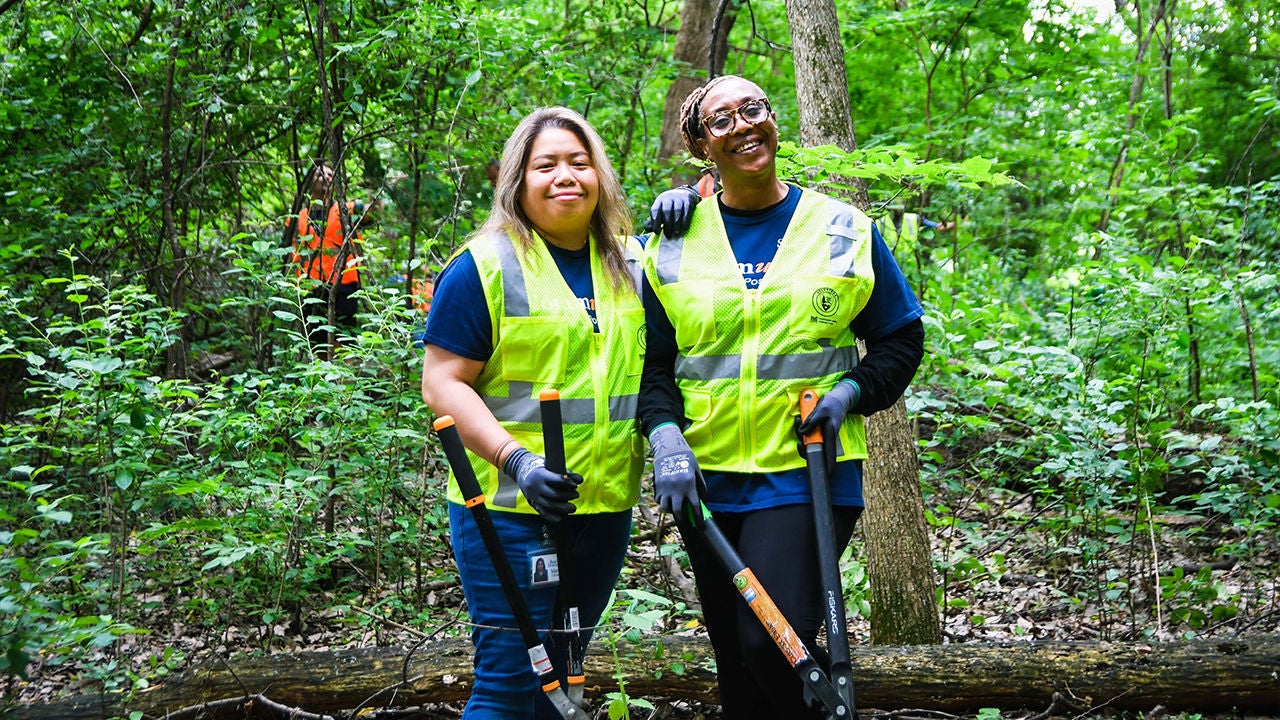 Two volunteers wearing high-visibility safety vests stand together in a lush green forest. 