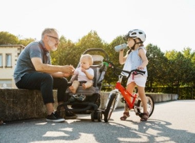 Grandfather sitting on curb with his young grandchildren in stroller and on bike.