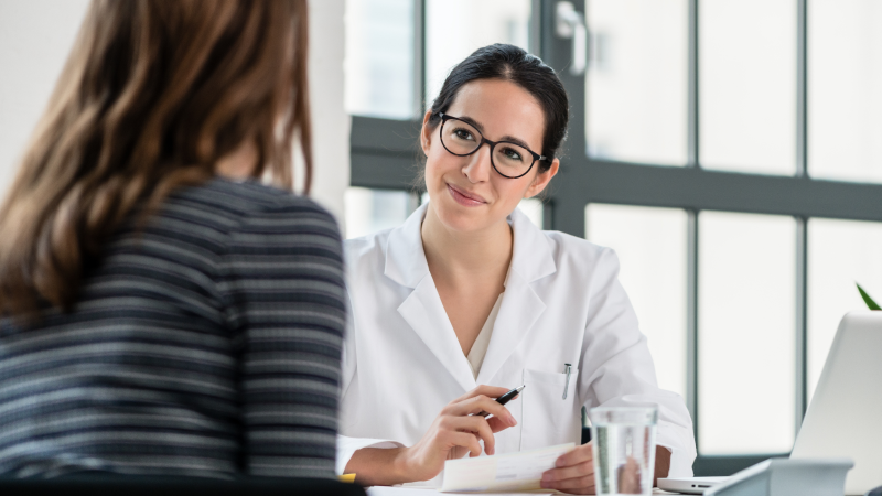 Health care professional discusses information with another adult across a desk in a clinical office.