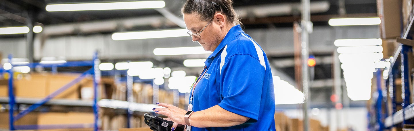 A warehouse employee in a blue uniform uses a handheld scanner while working in an industrial storage area. 