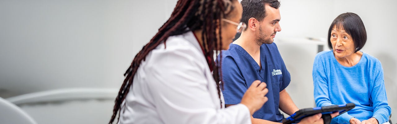 A healthcare professional in a white coat speaks with a patient and another clinician in a medical setting.