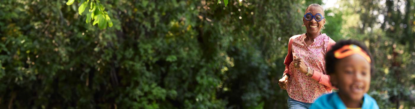Smiling woman wearing blue glasses and chasing boy through trees.