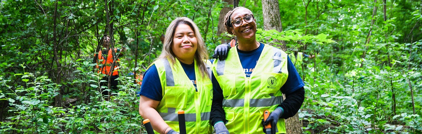 Two volunteers wearing high-visibility safety vests stand together in a lush green forest.