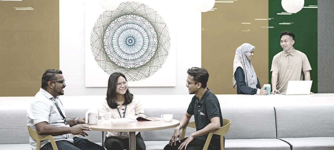 Three racially diverse employees sit together at a small table while a man and woman work together at a table behind them.