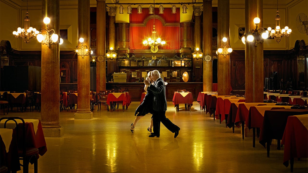 An elegantly dressed couple dances closely in the center of a spacious, ornate restaurant hall. Warm golden lighting reflects off the polished floor.