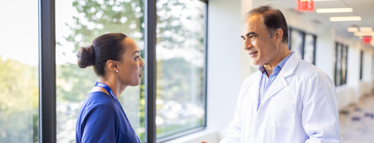 A woman wearing protective goggles and a hair covering looks into a microscope. 