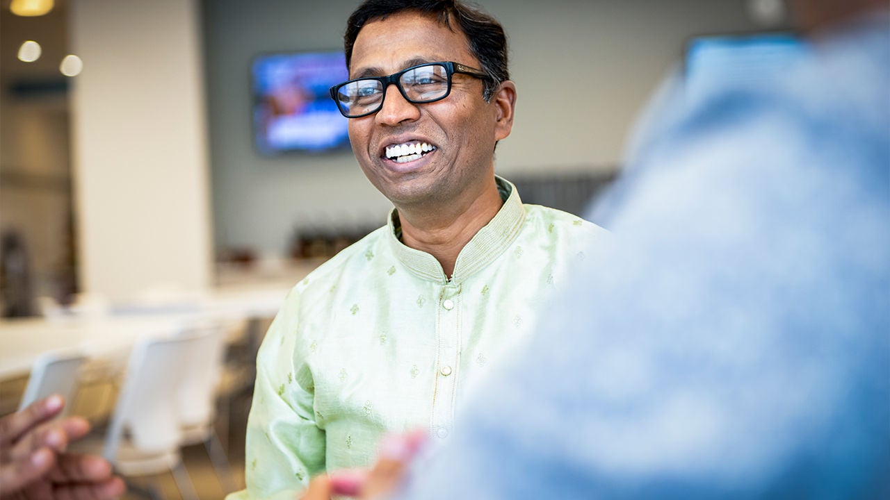 A man wearing a light green traditional-style shirt is engaged in conversation with others in an indoor setting. 