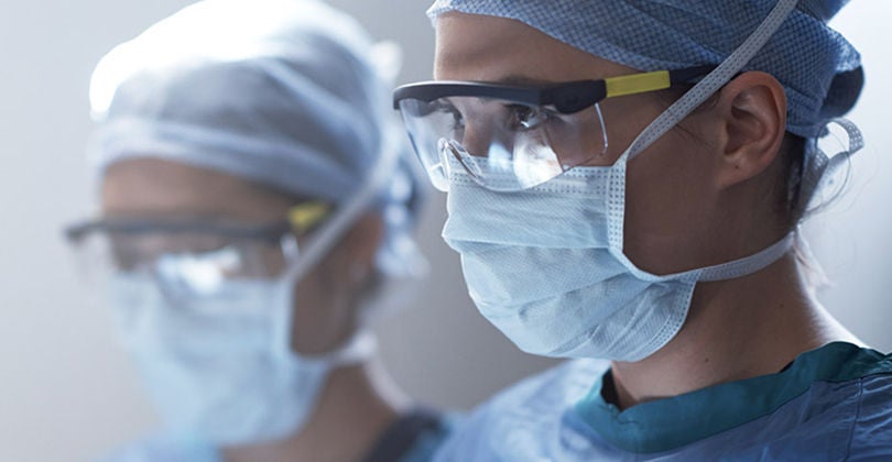 Close-up of two healthcare professionals wearing surgical masks, protective eyewear, and scrubs during a medical procedure.