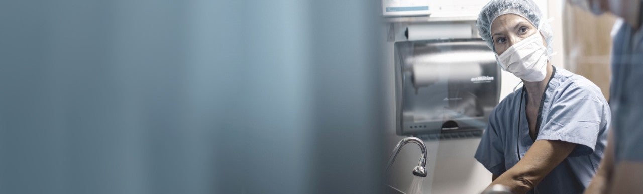 Healthcare worker in scrubs, mask and head cover washing hands and looking at colleague.