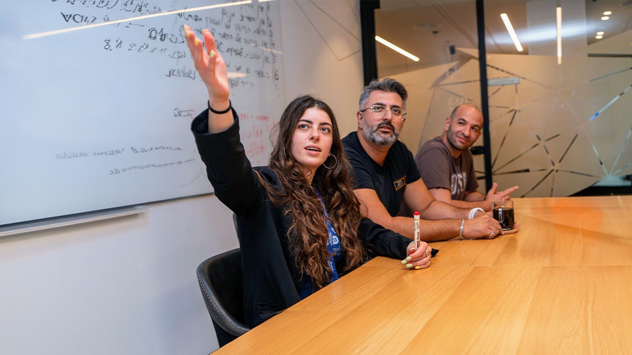 A smiling woman in an office environment points to a board that two male colleagues look at.