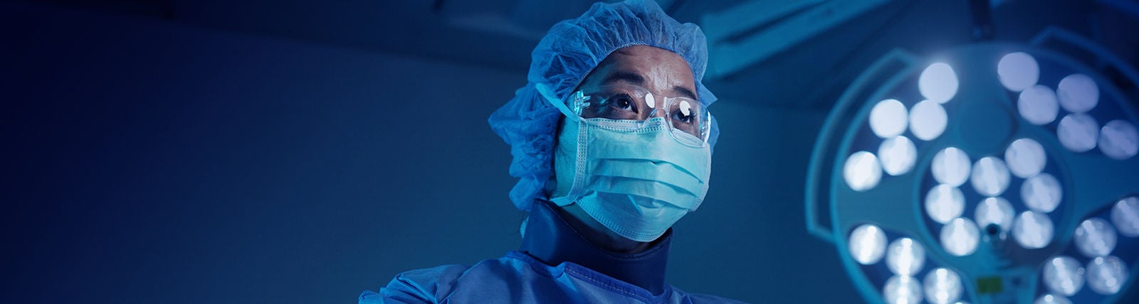 Surgeon in blue mask next to operating room lamp.