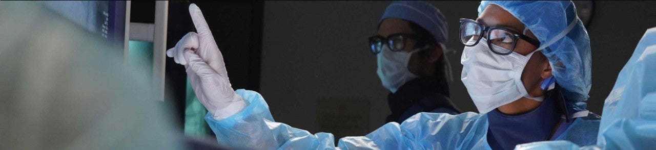 Medical professional in surgical gear pointing to screen as another looks on.