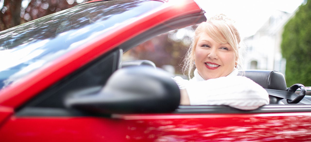 A smiling woman in a white sweater driving a red convertible with the roof open, enjoying a sunny day in a residential area.