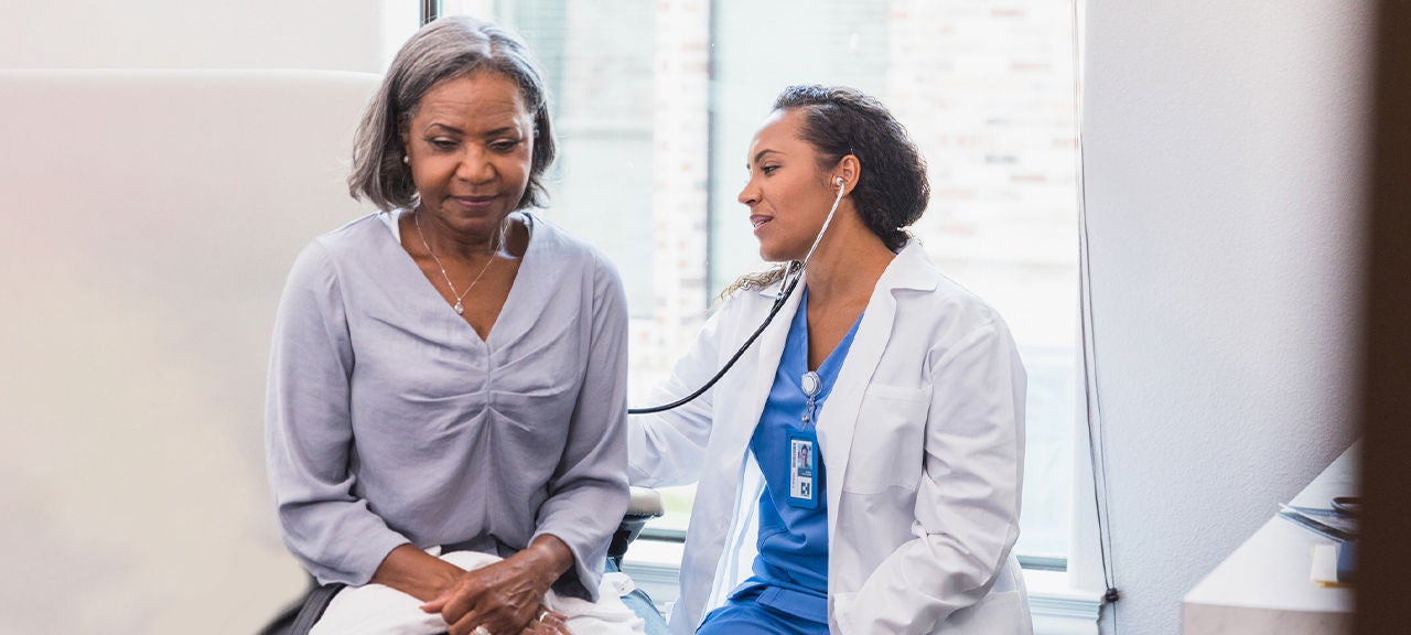 A female doctor wearing scrubs and a lab coat uses a stethoscope on the back of a female patient sitting on an examination table.