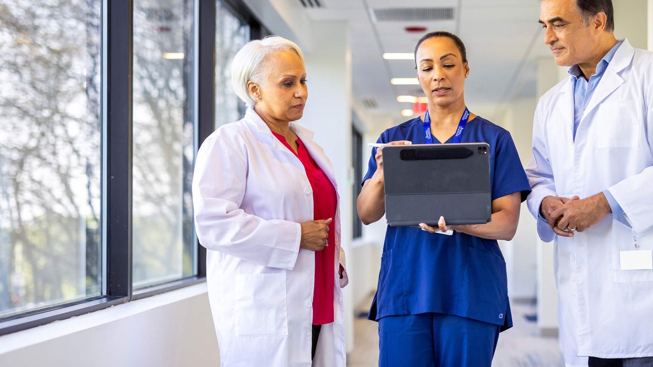 Three healthcare professionals standing in a hallway reviewing information on a tablet.