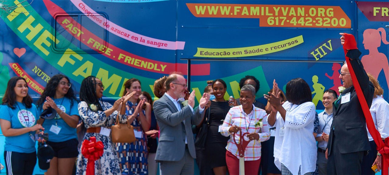 A diverse group of people attend a ribbon-cutting ceremony in front of a van that provides health care to underserved Boston communities.
