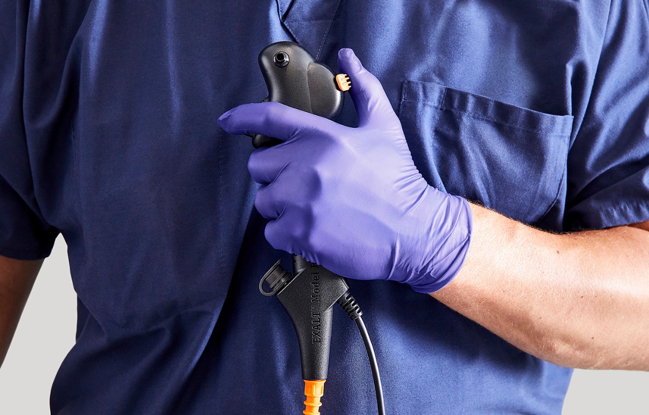 Medical provider's gloved hand holding EXALT Model B Single-Use Bronchoscope in front of their chest.