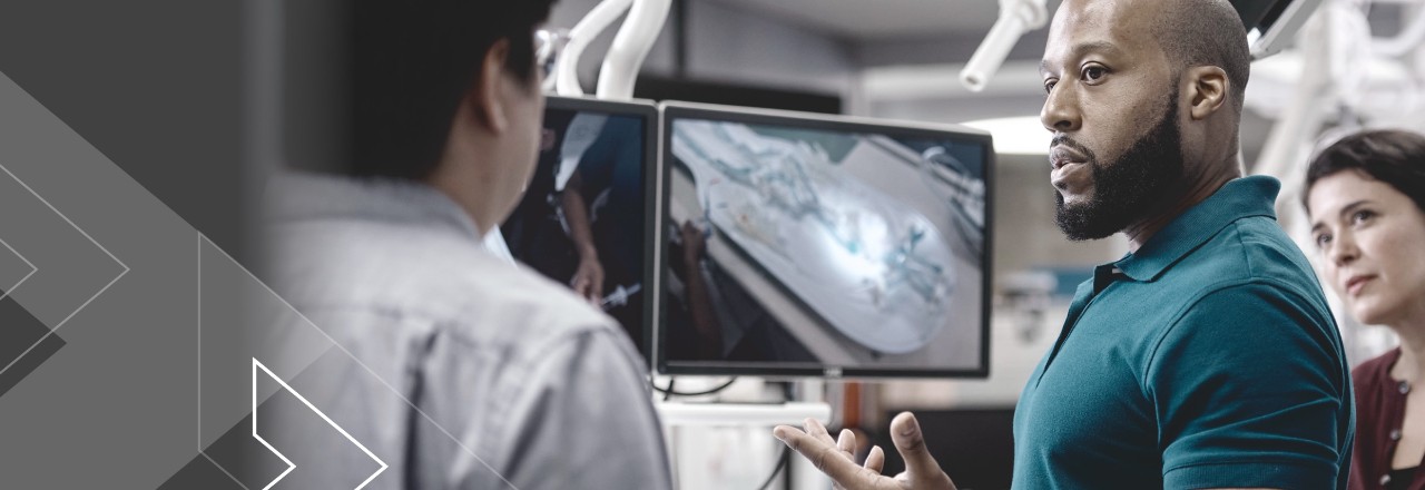 Man in front of monitor with image of medical device, gesturing to another person while woman looks on.