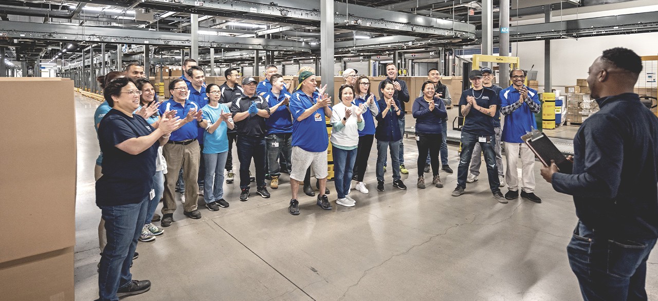 A group of Boston Scientific employees gather around a leader in a warehouse while clapping and cheering.