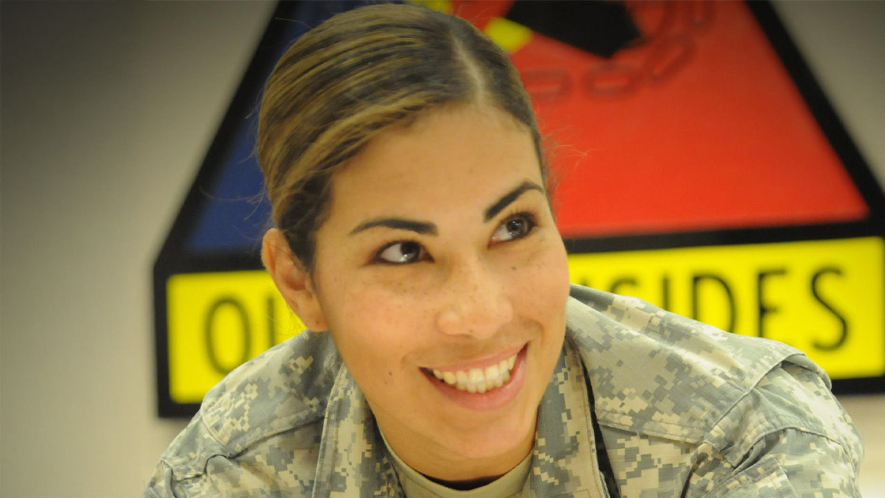Smiling woman in a U.S. Army camouflage uniform looking to the side, with a colorful background.