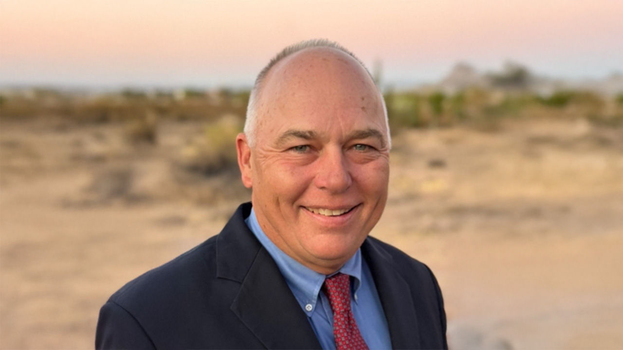 Portrait of a smiling man in a suit and red tie standing outdoors in a desert landscape.