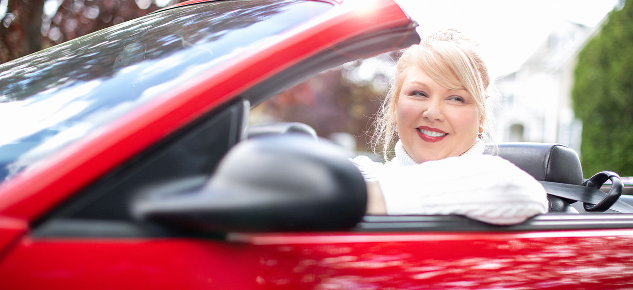 A smiling woman in a white sweater driving a red convertible with the roof open, enjoying a sunny day in a residential area.