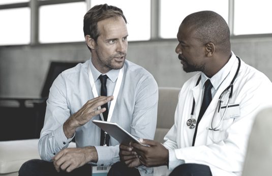 Seated medical sales rep speaking to seated doctor holding tablet device 