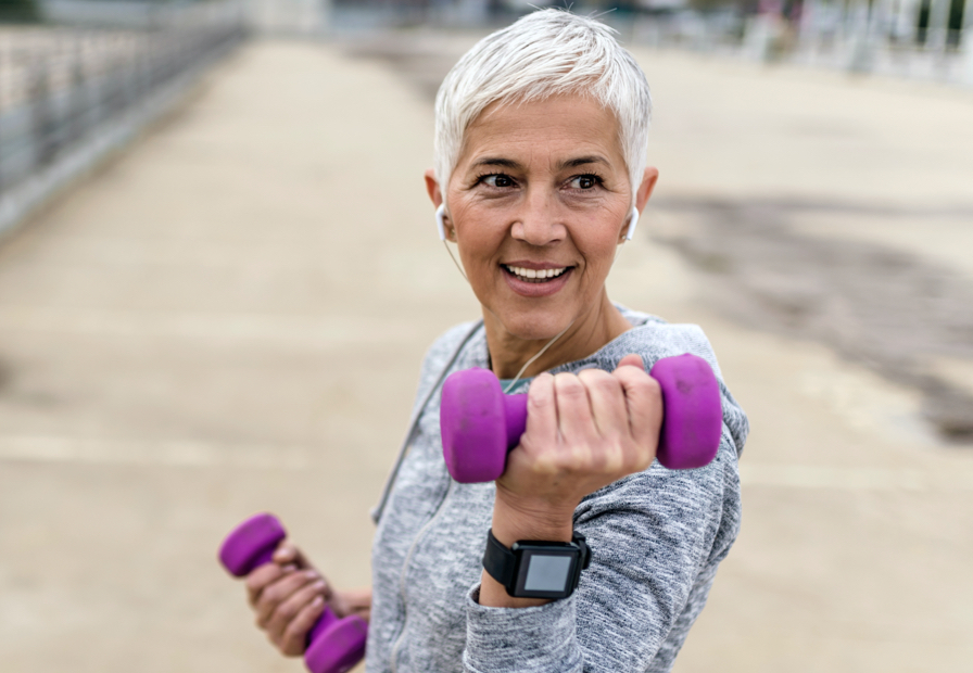 Woman smiling and lifting weights outside with headphones in.