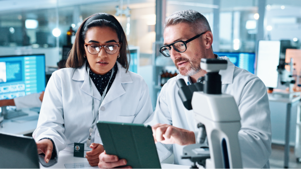 Two laboratory professionals review data on a tablet beside a microscope in a research lab.