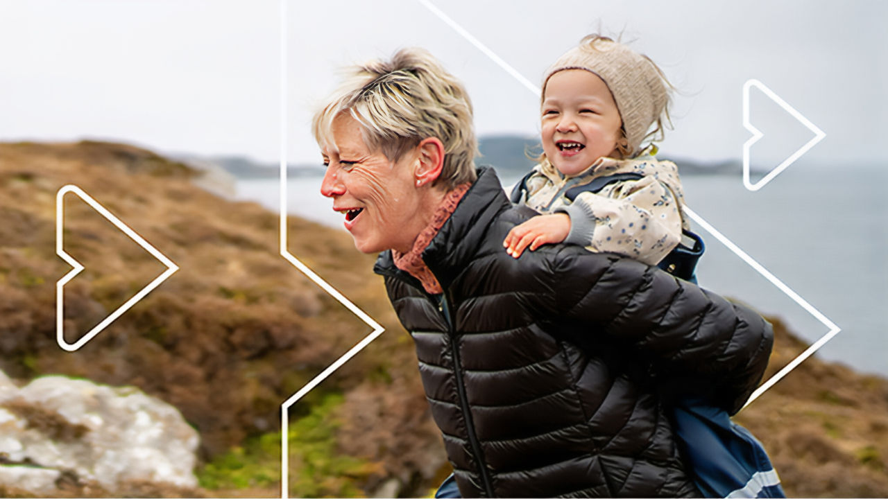 Smiling adult giving a child a piggyback ride along a coastal path.