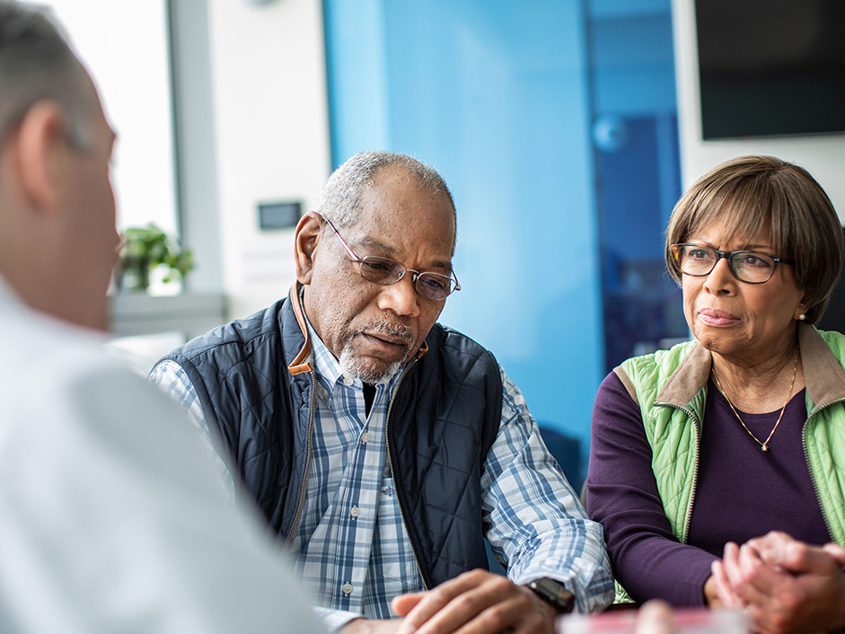 Older male patient and partner listening to a clinician during a consultation.