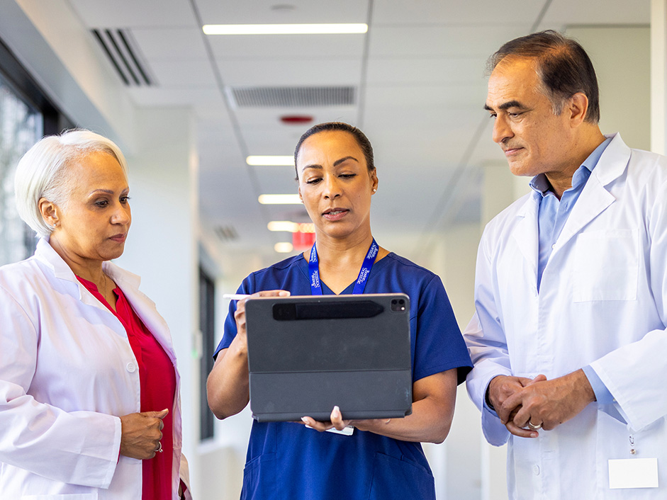 Three clinicians reviewing information on a tablet in a hospital corridor.