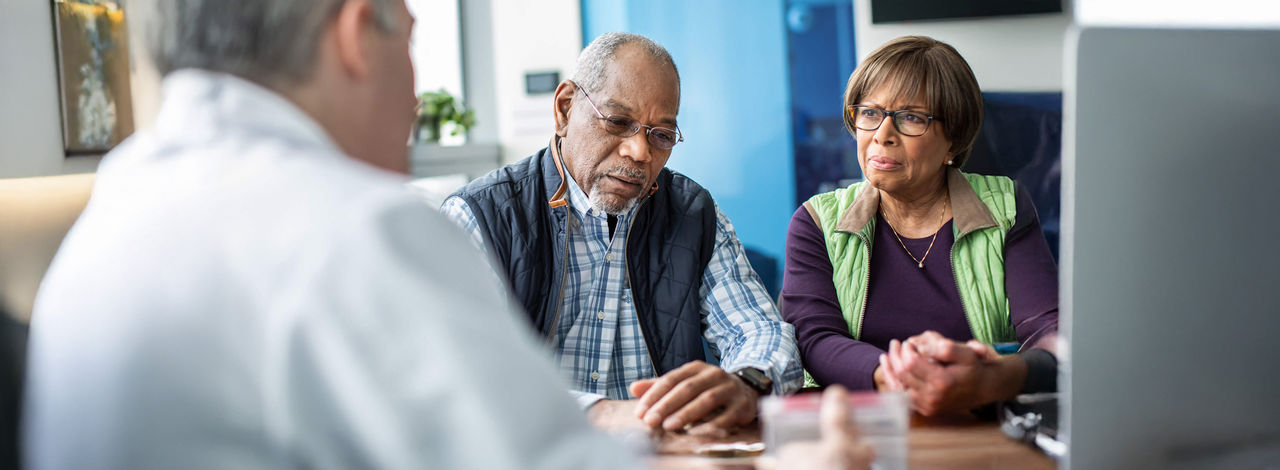 Older male patient and companion speaking with a clinician during a medical consultation.