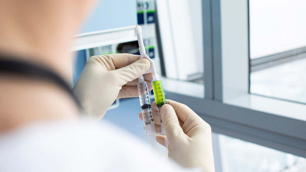 Gloved clinician holding connected syringes containing clear and green fluid in a clinical setting. 
