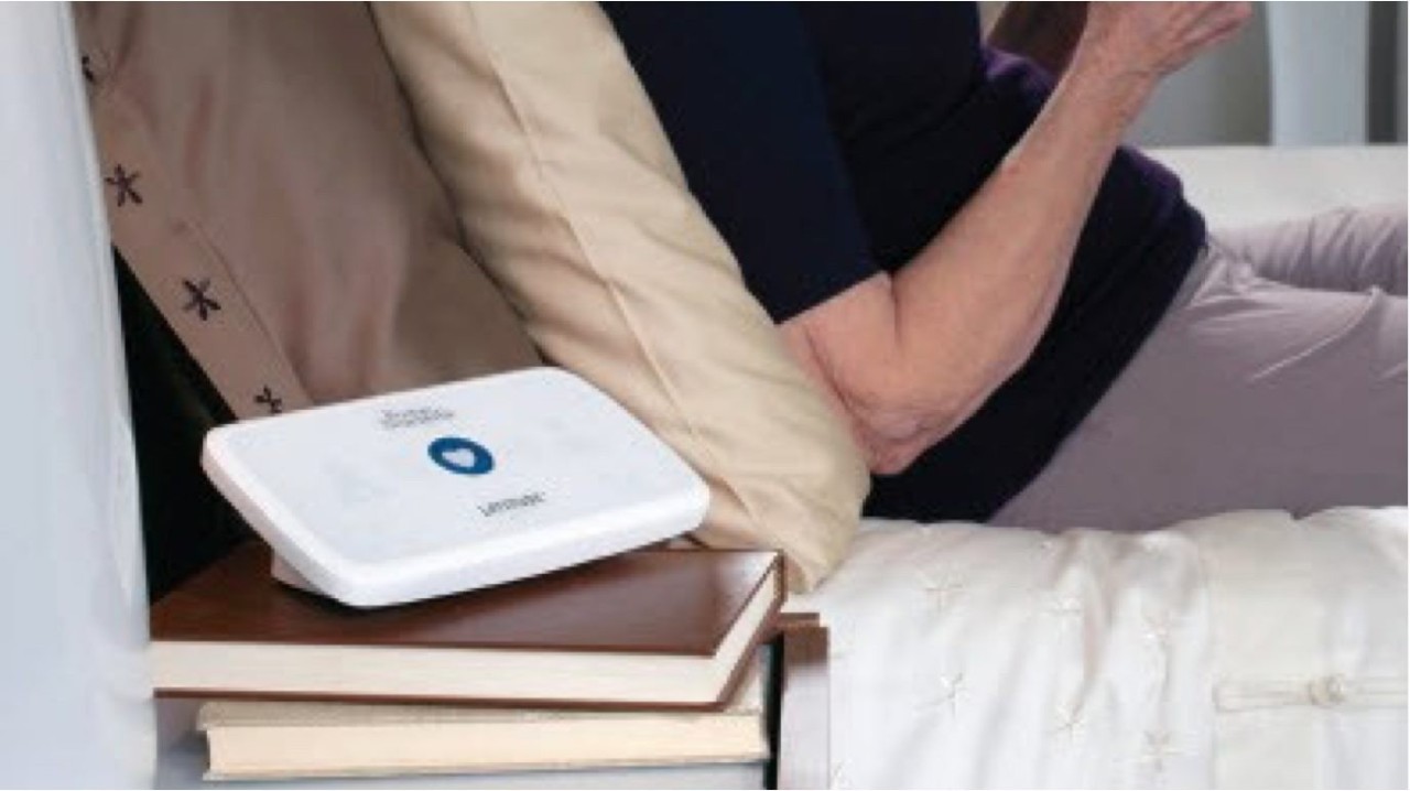 Boston Scientific’s LATITUDE Communicator sitting on a bedside table with a person lying in bed in the background